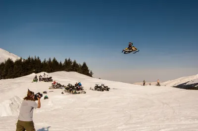 A high-action shot of a snowmobiler jumping through the air with others watching from a snowy ridge.